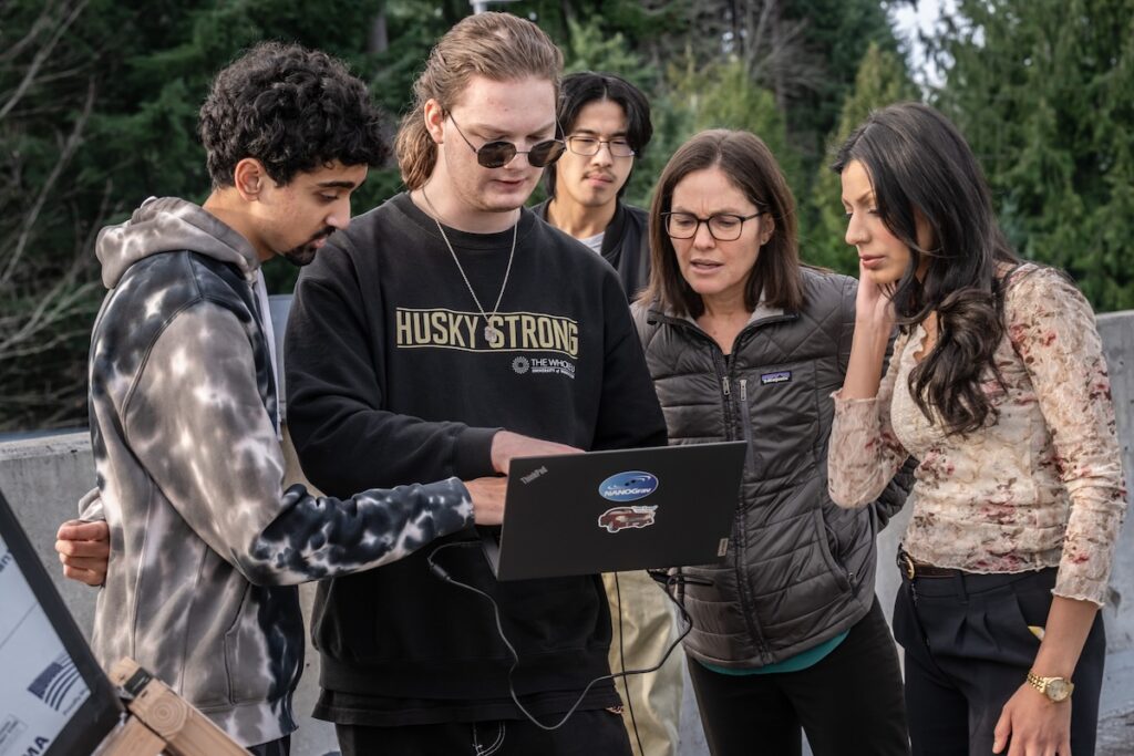 Five people collaborate outdoors while reviewing a laptop.