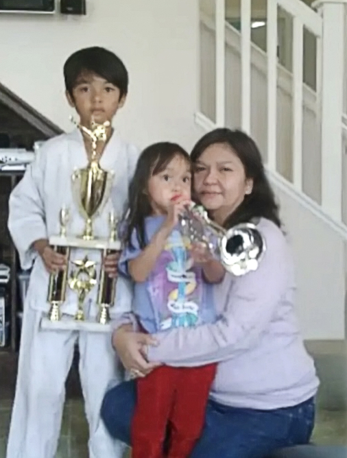Bilal, as a child, holding a martial arts trophy. Also in the photo are his younger sister, blowing a trumpet, and his mother kneeling down at the level of the kids. 