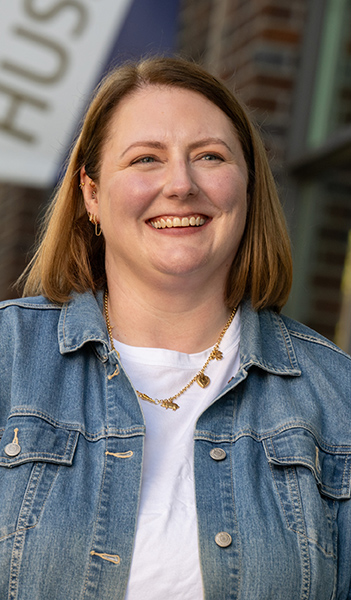Portrait of Nicole Sanderson in a denim jacket over a blue top. She's standing next to brick buildings with a slightly blurry "Huskies" banner behind her. She's laughing and looking off-camera.