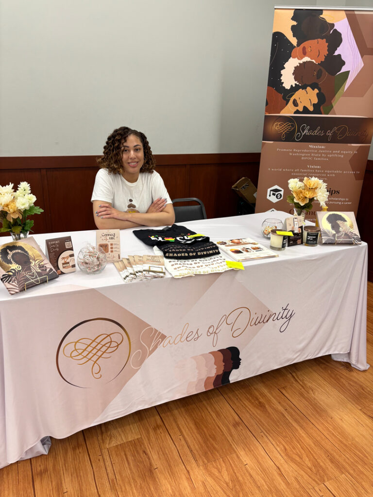Shanell Brown sits behind a Shades of Divinity information table displaying educational materials, community resources and branded items during a community event.