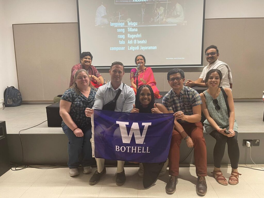 A group of people posing with a classic Indian music band on a stage while holding a University of Washington Bothell flag