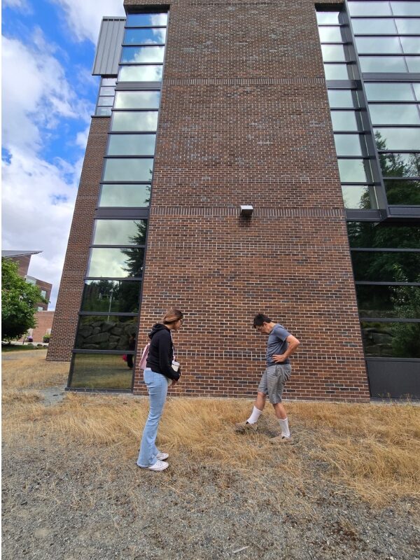 Salina and Dr. Jensen are standing and observing the site next to tall building with mirrored windows.