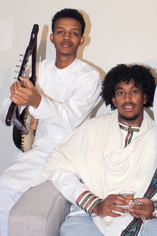 Tadesse and Legesse in traditional attire. Tadesse is holding a guitar-like instrument called a krar, and Legesse is holding a small cup of coffee.