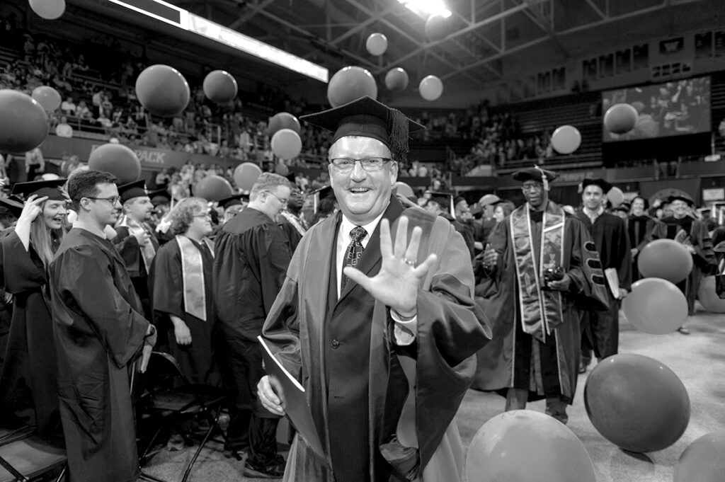 Bradley Portin, in full academic regalia, walking down the aisle while waving during commencement.