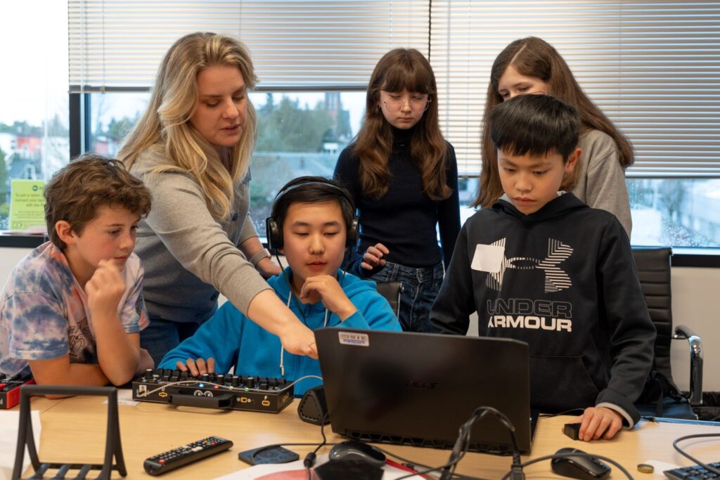 children stand around a laptop being taught by a teacher