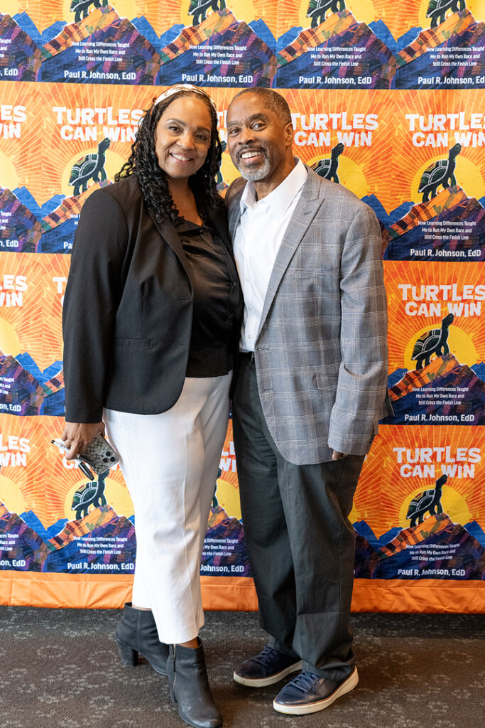 Paul Johnson and his wife, Ricole, smile together in front of a backdrop featuring the cover of his memoir, “Turtles Can Win.”