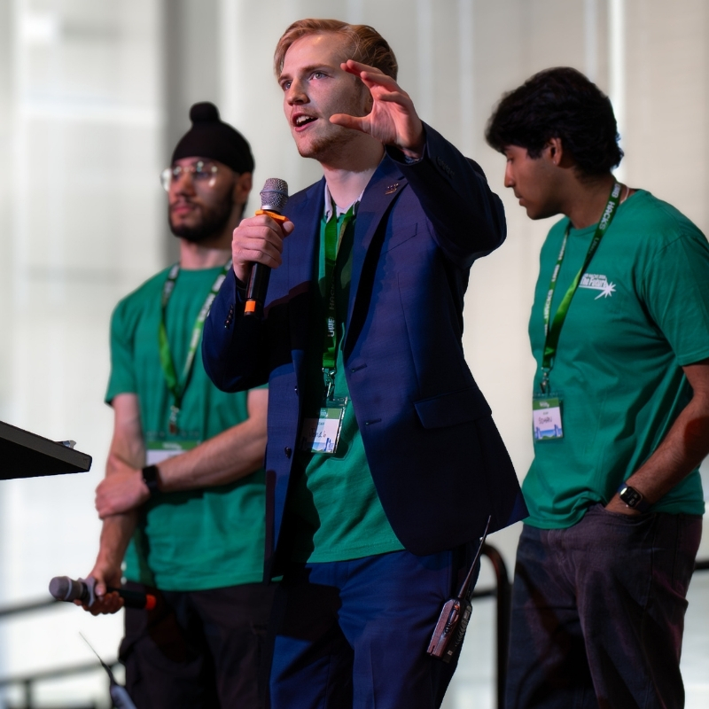 Jonah Uellenberg gestures while speaking into a microphone alongside two fellow participants at the 2026 Hackathon.