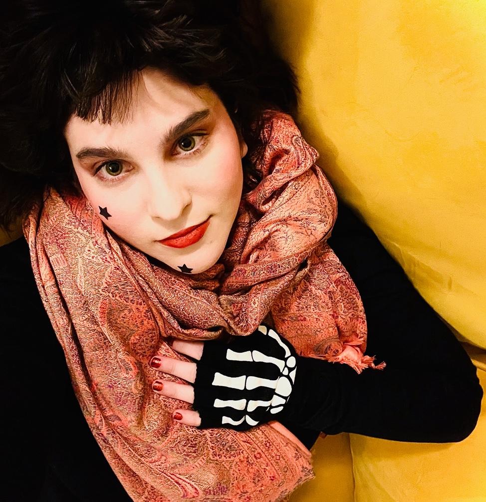 A.V. Marraccini poses in front of a yellow background. They are wearing black star stickers on their face and skeleton hand gloves.