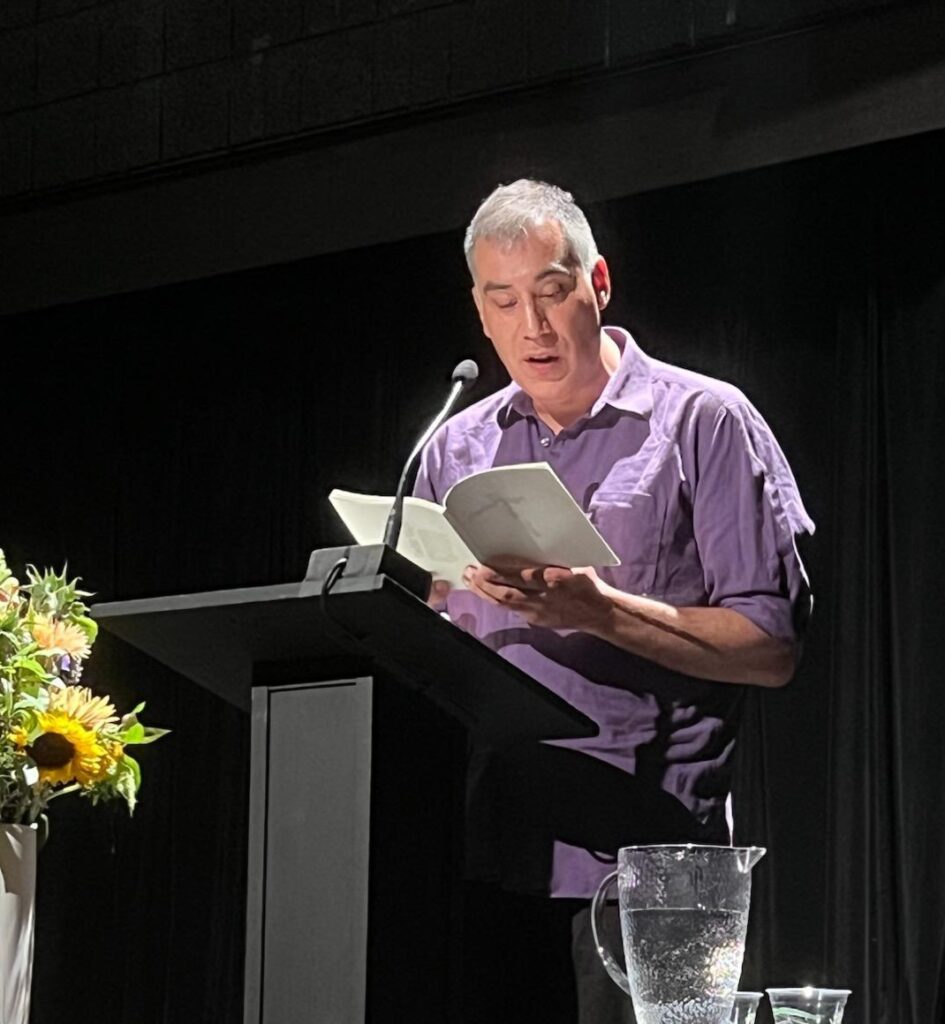 Cedar Sigo is wearing a purple shirt and reading from a book on stage at an event in from of a black curtain.