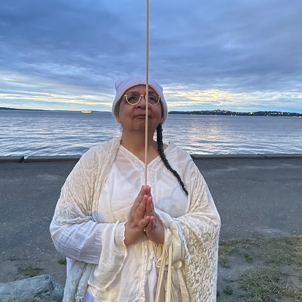 Nisi is hearing a white dress and cream shawl. They are on coastal beach holding a wooden stick.