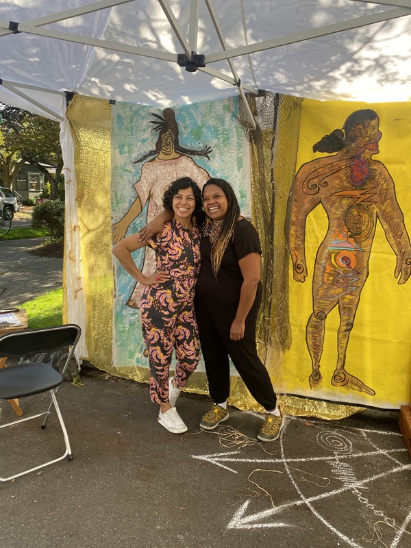 Photograph of two women standing outdoors under a canopy tent. Behind them are hung tapestries featuring colorful art images. 