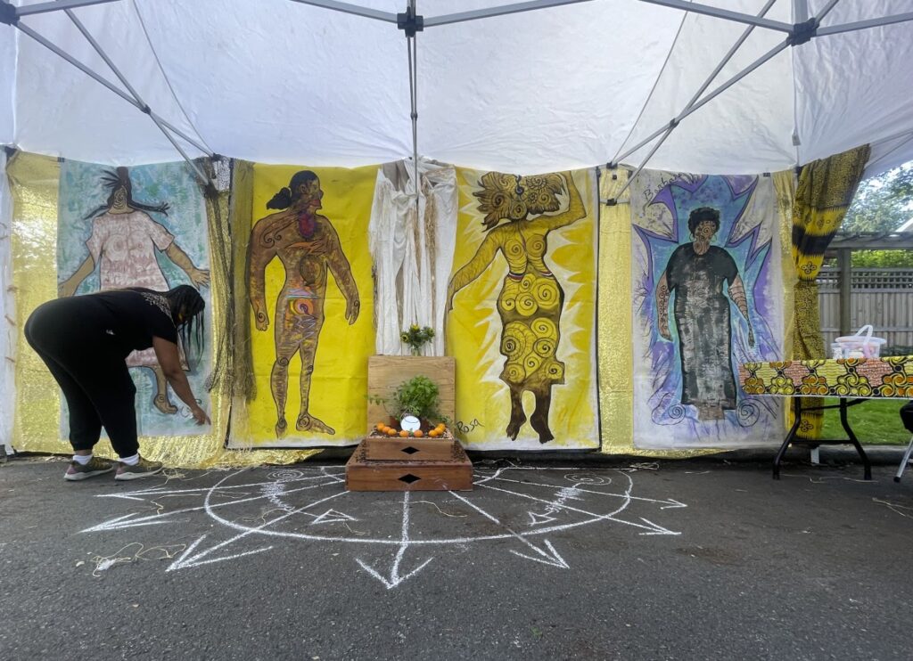 Photograph of asphalt road with chalk drawings. A canopy tent is overhead and colorful hanging tapestries are hung along backside of tent. A woman is crouched near bottom of tapestry.