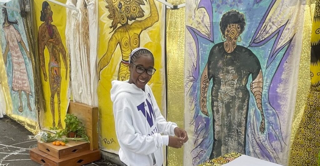 A person wearing a white UW Bothell sweatshirt standing in front of colorful hanging tapestries.