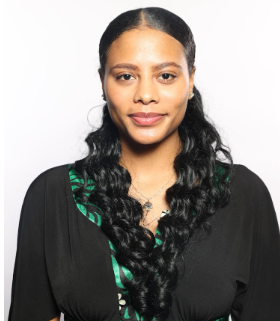 A woman with long, dark, wavy hair styled in two sections looks directly at the camera with a composed expression. She is wearing a black top with green patterned accents and small stud earrings, with a simple necklace layered around their neck. The background is plain white.
