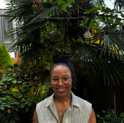 Asia smiles in front of a palm tree backdrop, wearing a white stripped top.