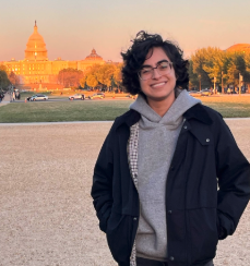Damien stands outdoors under soft light infront the US White House, wearing glasses and a dark jacket, looking directly at the camera.