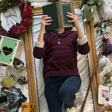 Mason laying reading a book on a white cloth and within large golden frame. The background surrounding them contains the placement of different whismical objects.