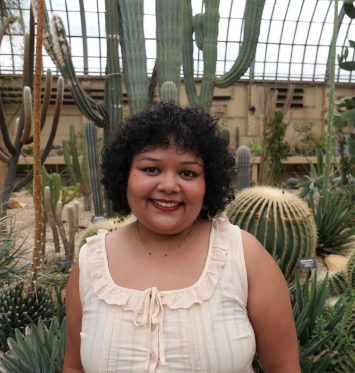 Noor stands in front of green cacti of different sizes and hues of green, wearing a light cream shirt with fringe.