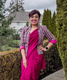  woman standing outdoors smiling confidently, wearing a magenta sari paired with a patterned blazer that has rolled-up sleeves. She has short hair, silver earrings, and a smartwatch. Evergreen trees and suburban houses appear in the background under a cloudy sky.