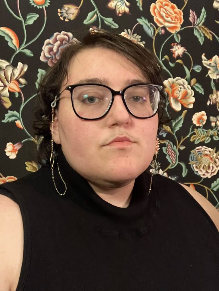 A person with short brown hair and glasses looks directly at the camera with a calm expression. They are wearing a sleeveless black turtleneck and long, asymmetrical dangling earrings. The background features a richly patterned floral wallpaper with large blossoms in muted tones.
