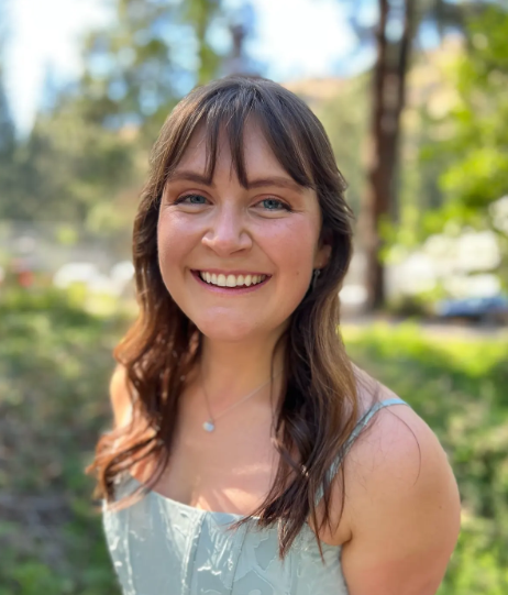 Liz Daniel smiles in front of a blurred outdoor background with green foliage. She has shoulder-length brown hair and is wearing a mint green dress.