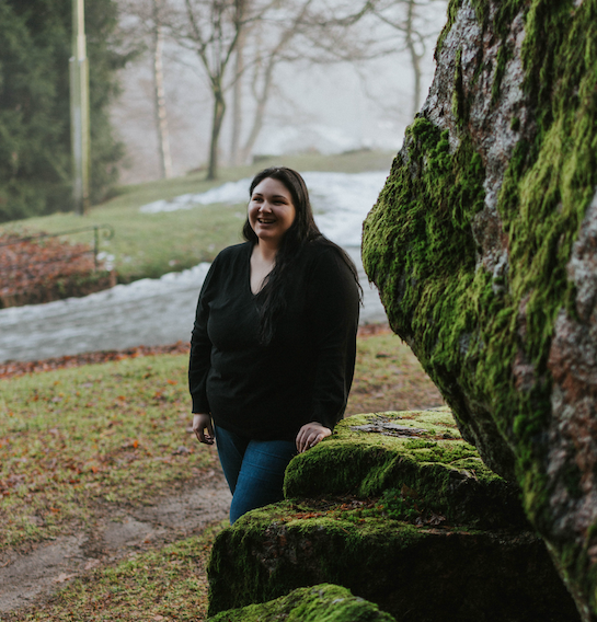 Angelica smiling, wearing a oversized black sweater. She stands by a large boulder covered in moss.