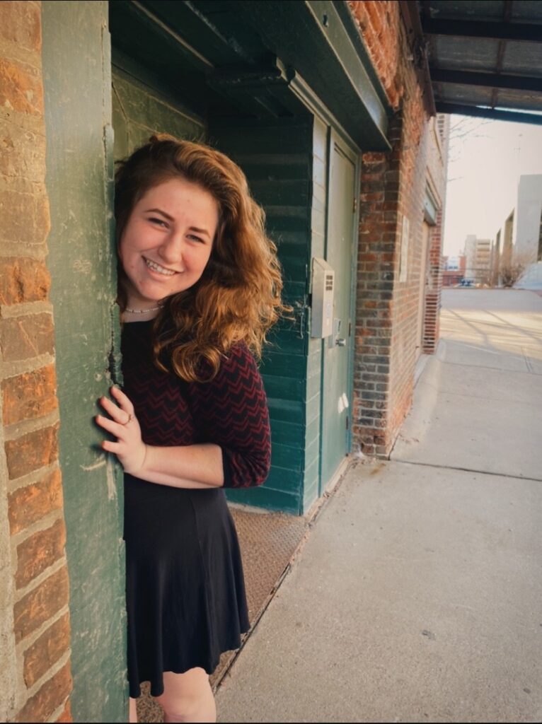 A woman with long, wavy brown hair smiles brightly while leaning playfully from behind a green wooden doorway. She is wearing a maroon and black patterned top and a beaded choker necklace. 