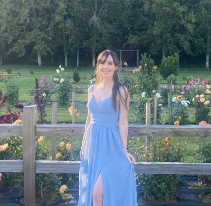 Felicia Zsiros smiles while wearing a periwinkle long dress. She is standing against a wooden fence and a flower garden of various colors.