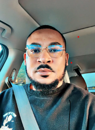 Emmanuel Kofi Dei Ahenkorah wearing glasses and a gray sweater with a white logo on the left. He is standing against a light gray background with soft lighting in a car.