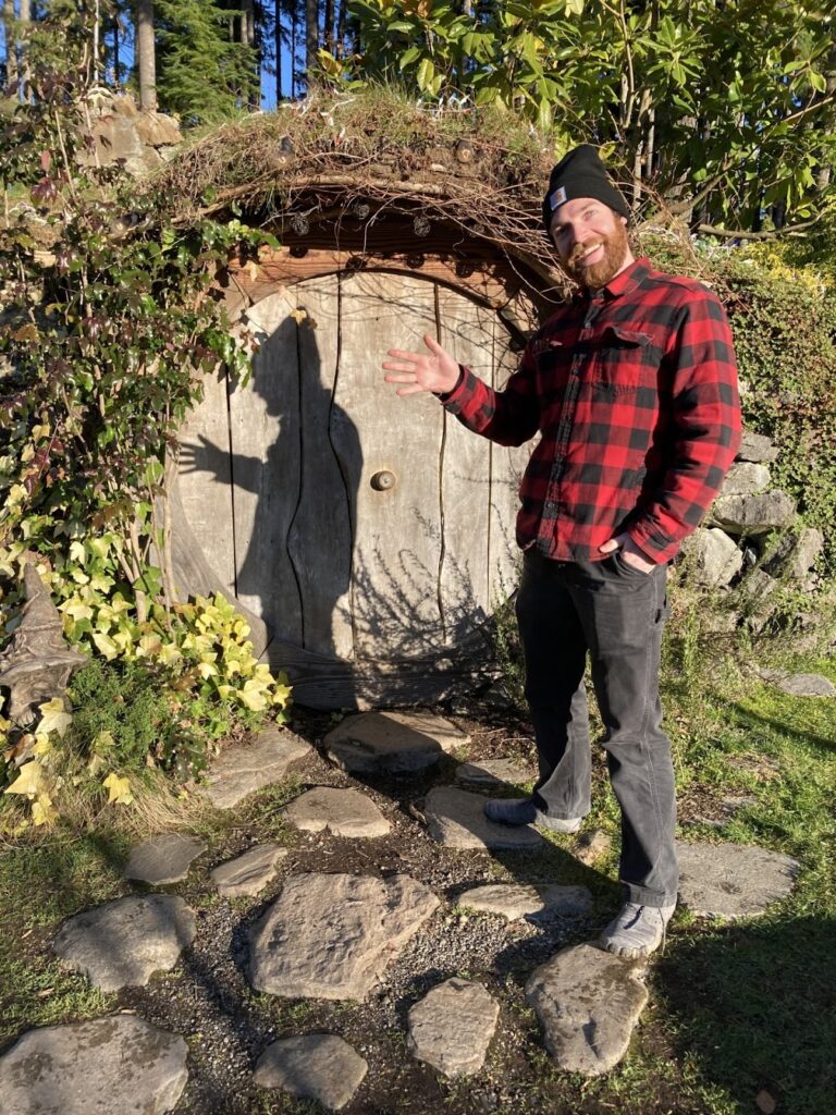A man with a beard smiles warmly while gesturing with one hand toward a round wooden door set into a stone and vine-covered wall. He is wearing a red and black plaid flannel shirt, black pants, and a black beanie. 