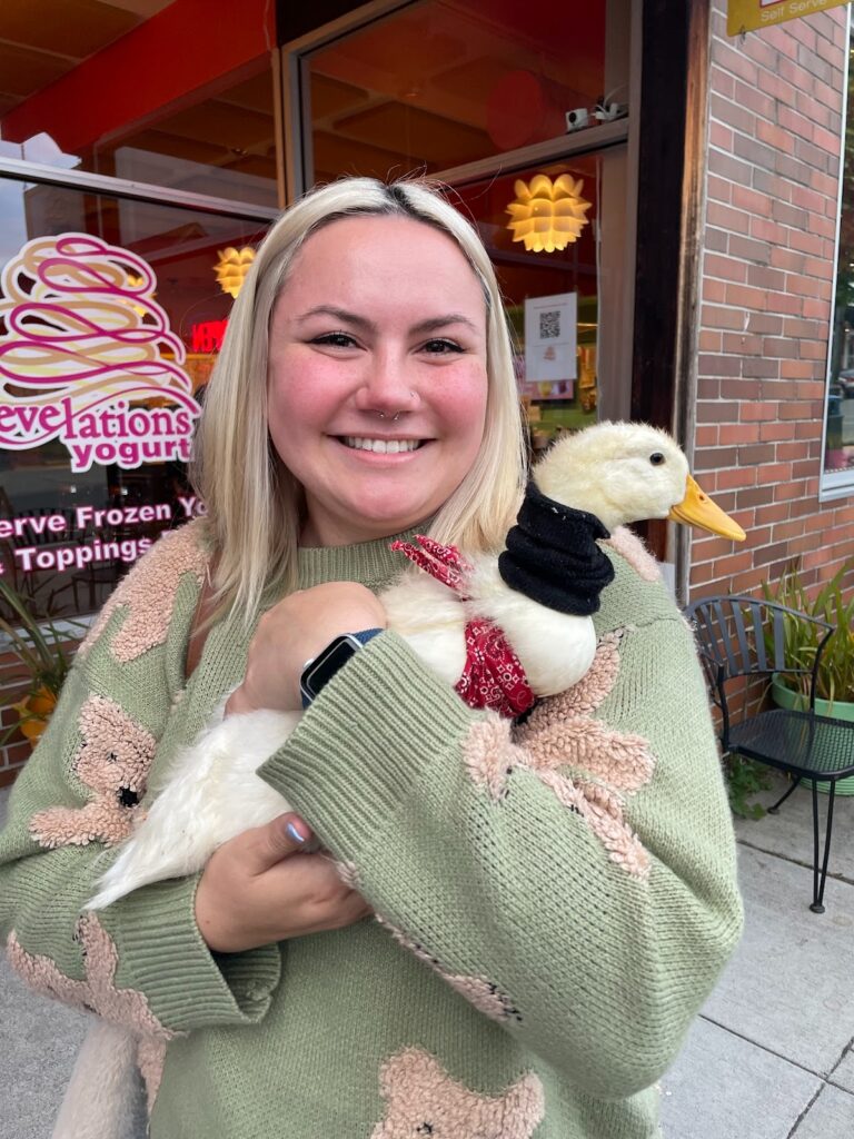 A woman with straight blonde hair smiles while holding a white duck wearing a small red bandana and black sweater. She is dressed in a sage green sweater with floral patterns and standing outside a yogurt shop with a colorful sign reading “Revelations Yogurt.” 