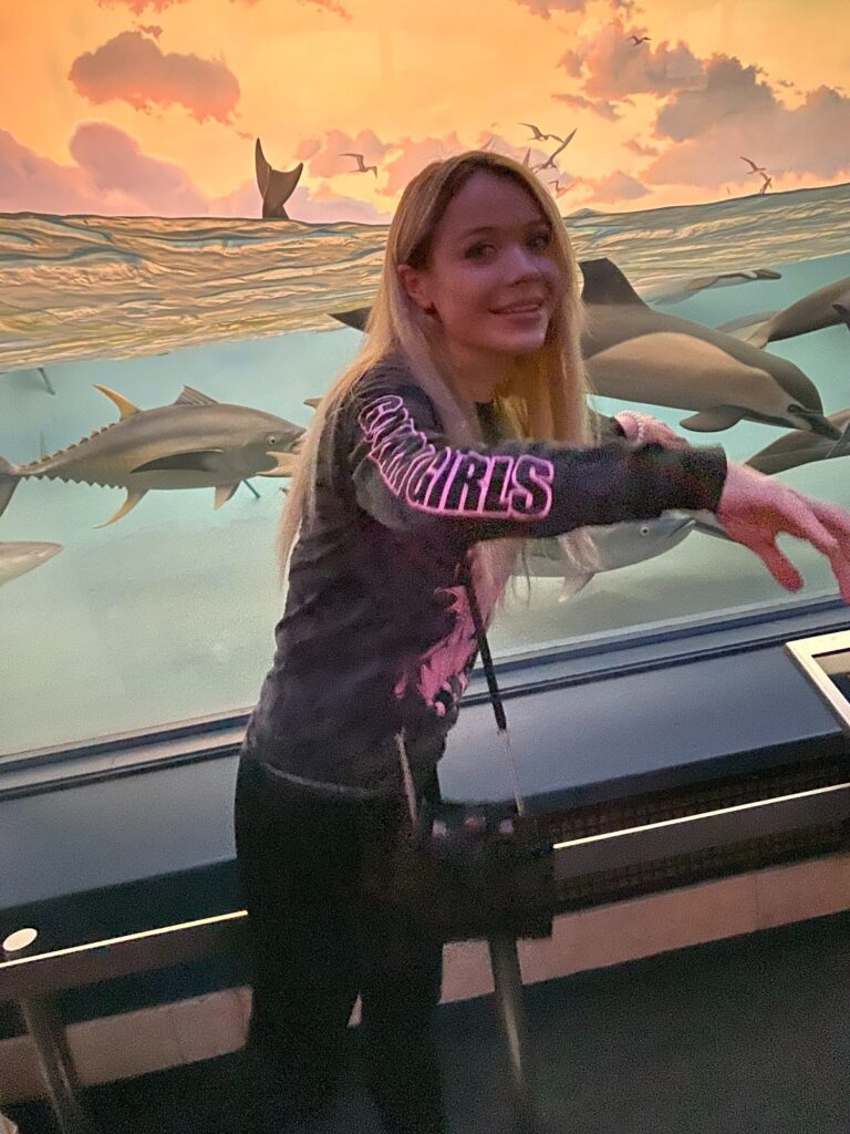 A woman with long blonde hair smiles while leaning slightly toward the camera, standing in front of a large aquarium display featuring swimming sharks and a sunset-lit ocean backdrop. She is wearing a black sweatshirt with pink lettering on the sleeve that reads “COWGIRLS.” 
