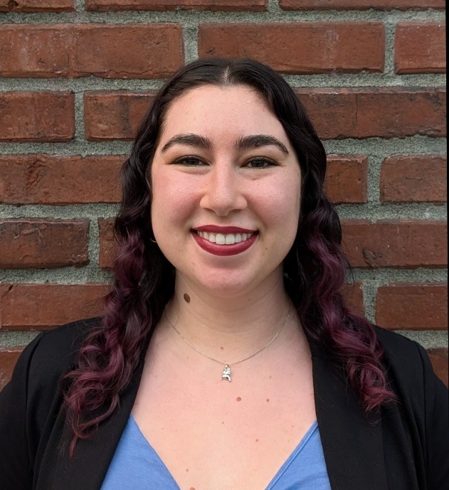 A woman with long, dark, wavy hair that fades into purple at the ends smiles warmly while standing in front of a brick wall. She is wearing a light blue top with a black blazer and a small silver necklace. 