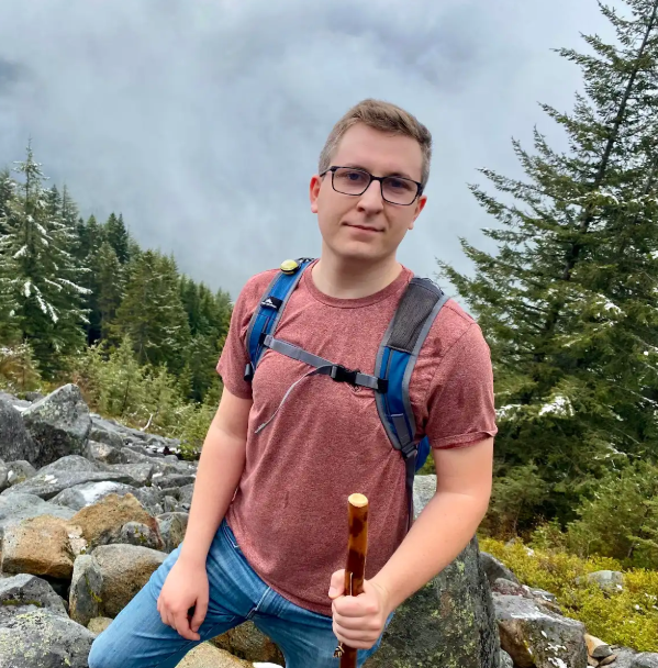 Ben Stafford smiles while wearing a muted red tshirt. He is photographed outdoors with rocks and several pine trees in the background.