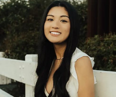 Anna Templeton smiles in front of a white wooden fence with green foliage in the back. She is wearing a white blouse and has long black hair.