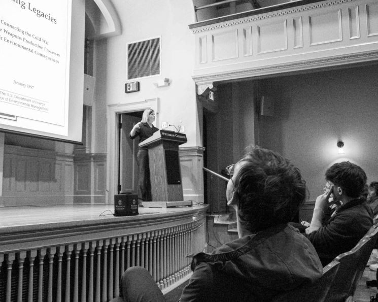 Black and white photograph of Cram standing on a stage behind lecturn. Cram is talking to an audience, also pictured.