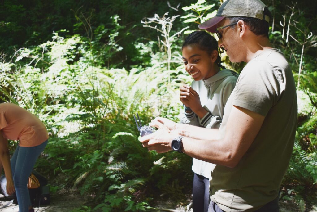 Two people in a forest area, one holding a remote control.