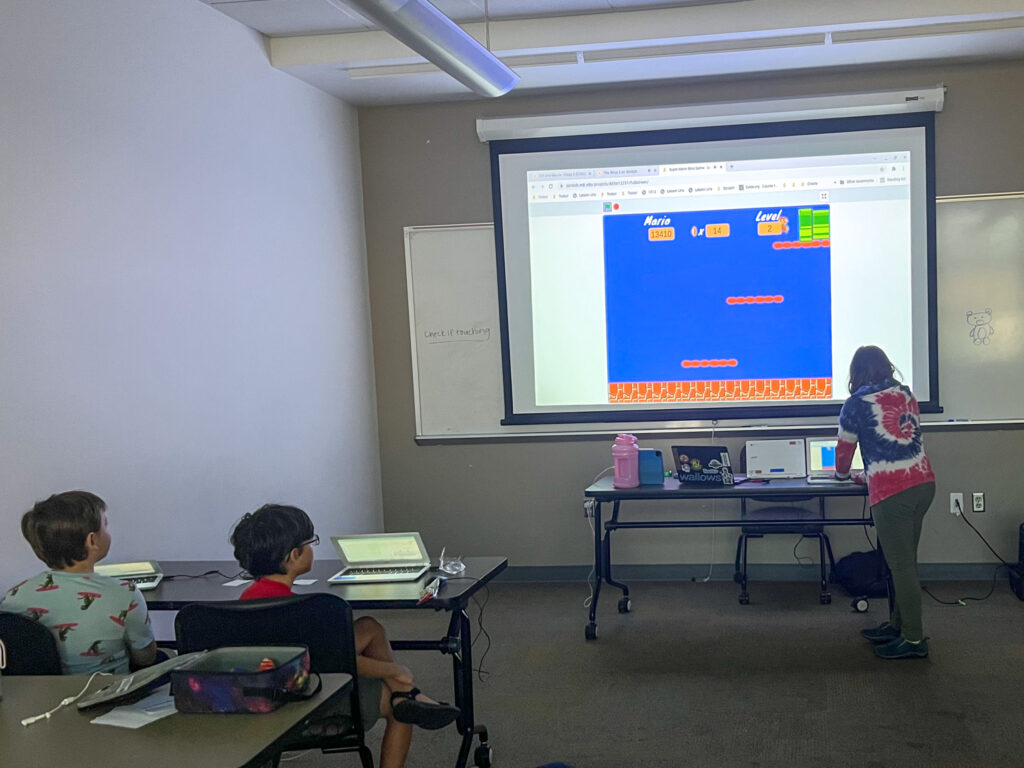 A person at a computer projecting a game on a screen with two children at a desk nearby.