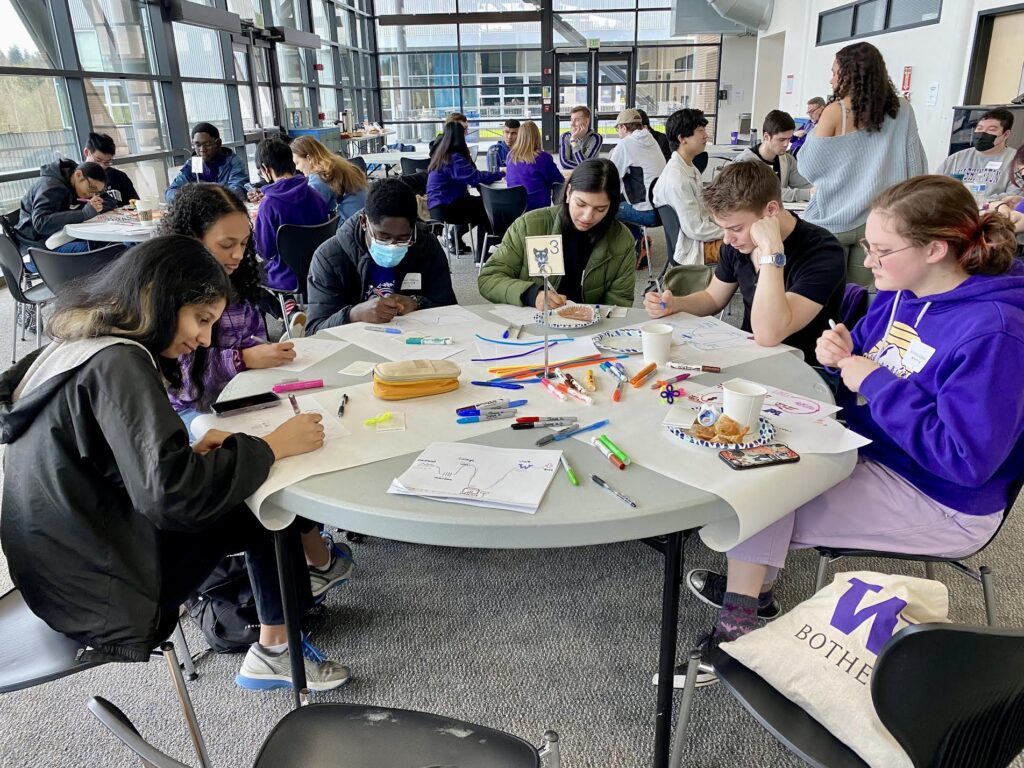 Groups of people sitting at tables writing and drawing on posters.