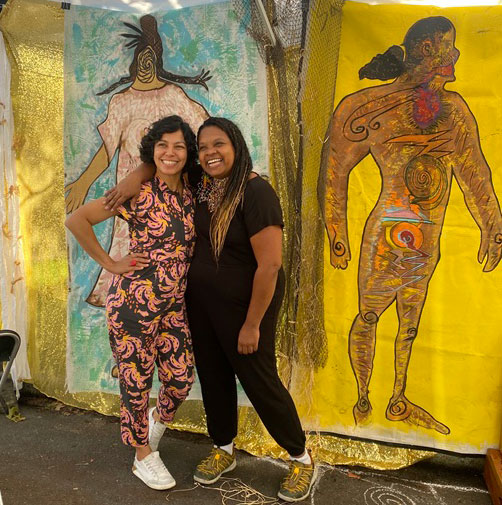 Two people pose for a photo in front of an art piece.