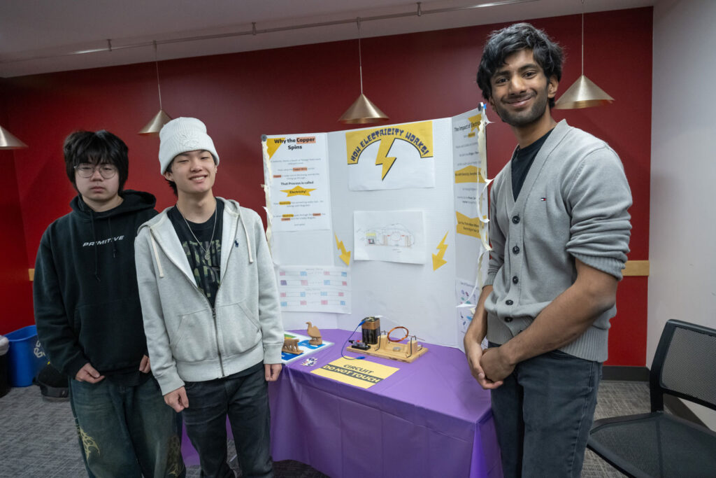 Three people stand next to a poster about electricity.
