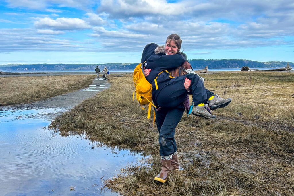 A person in waders carries another person across water.