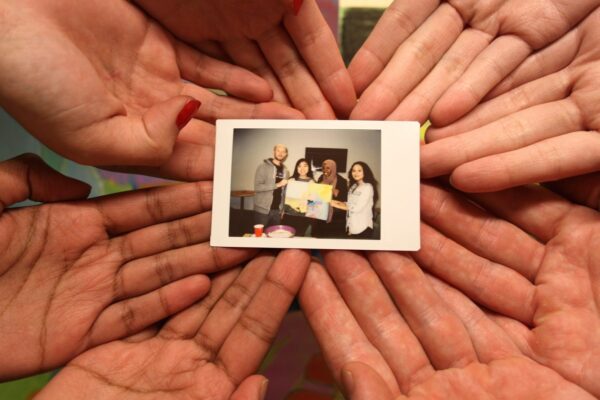 Multiple pairs of hands holding up a polaroid of four people holding a piece of artwork.