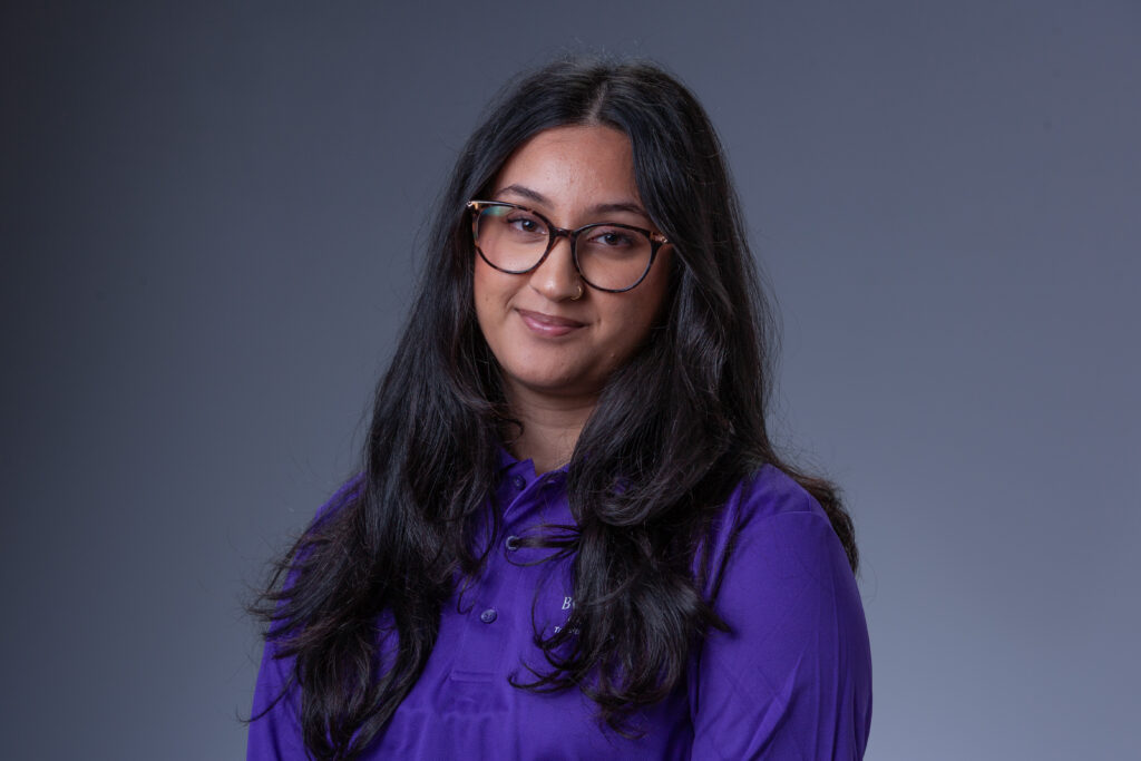 Peer Coach Reya in a purple polo in front of a gray background