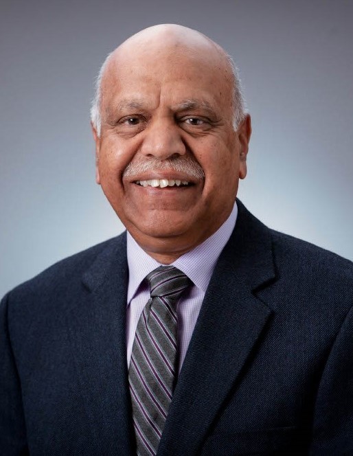 Dr. Gowri Shankar smiling in a dark blue suit, lilac collard button up, and a striped grey and dark purple tie.