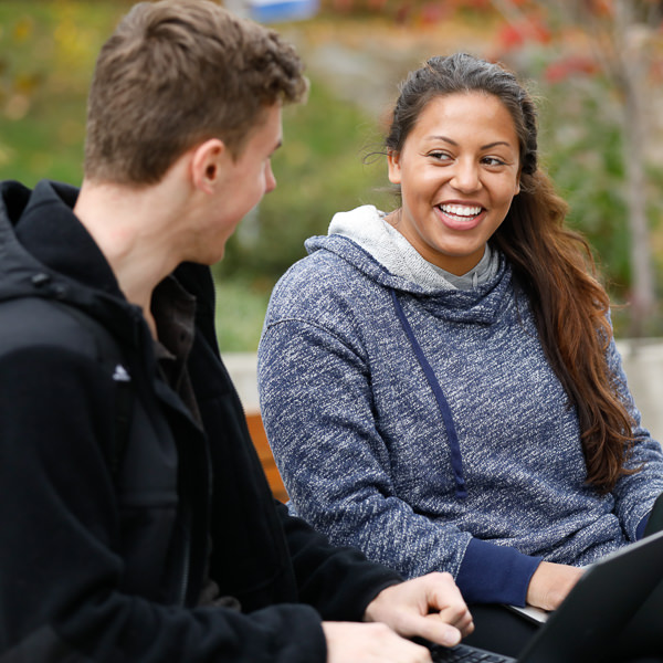 Female with dark hair in a gray hoodie with a computer looking at a man with brown hair wearing a black jacket.