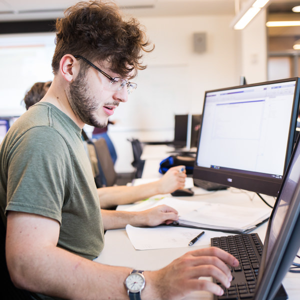 Man with dark curly hair with glasses wearing a green shirt working on a computer.