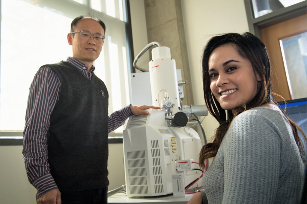 Dr. Choi standing next to Malia Steward who is sitting in front of the scanning electron microscope.