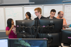 faculty and students standing behind a bank of monitors in a computer science classroom
