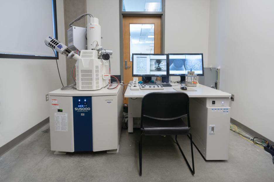 A room with a Field Emission Scanning Electron Microscope on the left, and a table with two monitors, a keyboard, mouse, chair and various equipment on the right. The monitors show microscope data.